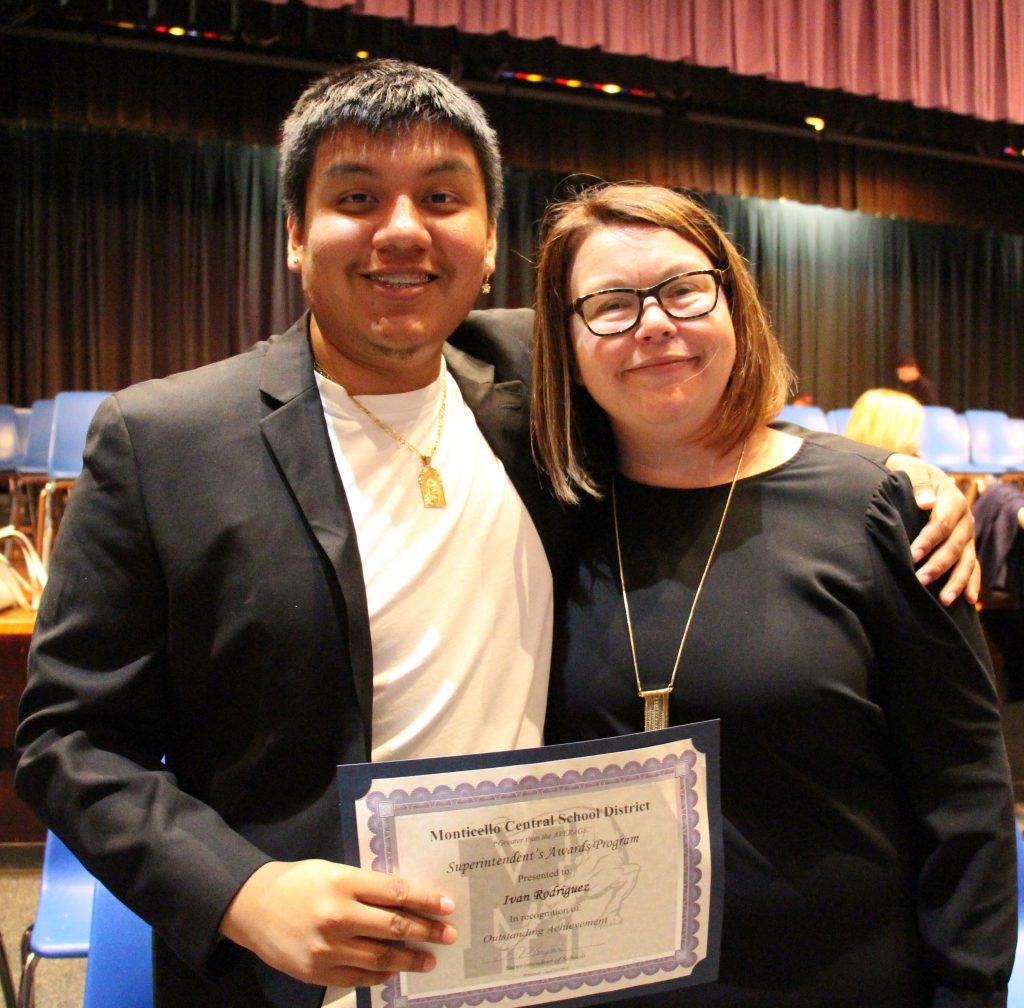 Young man with short dark hair, black suit jacket and white shirt holds a certificate in front of him. He has his arm around a woman with glasses and shoulder length hair wearing a black dress.