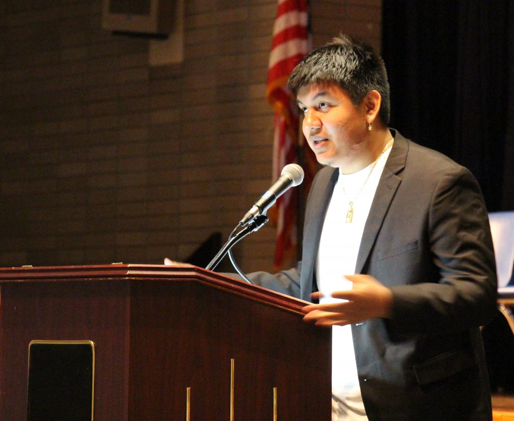 Young man in a black suit jacket and white shirt speaking at a podium.