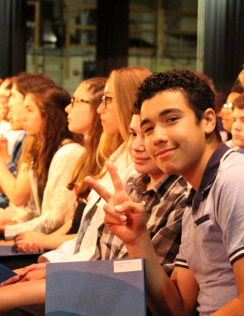 A row of students with one young man on the end holding his certificate and giving the peace sign.