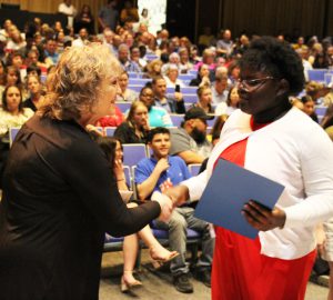 Woman in a black sweater shakes hands with a high school student wearing a red dress and white sweater. She is holding a certificate in her other hand.