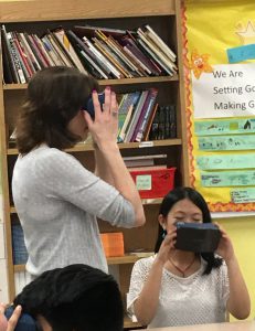 A teacher standing holding virtual reality goggles while two students sit looking through theirs
