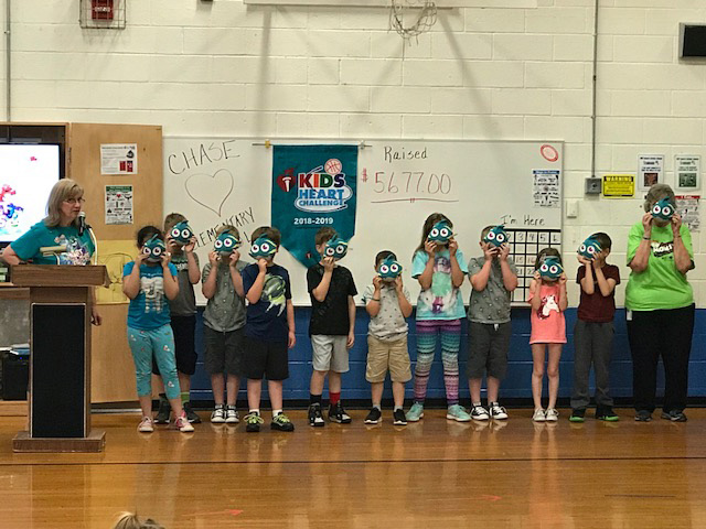 A line of elementary school students wearing masks stand in front of a sign showing how much money they raised for the Kids Heart Challenge.