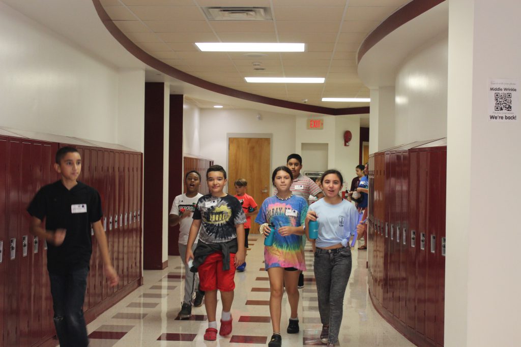 A group of new sixth-grade students walk down the hall. Red lockers are on either side of them.
