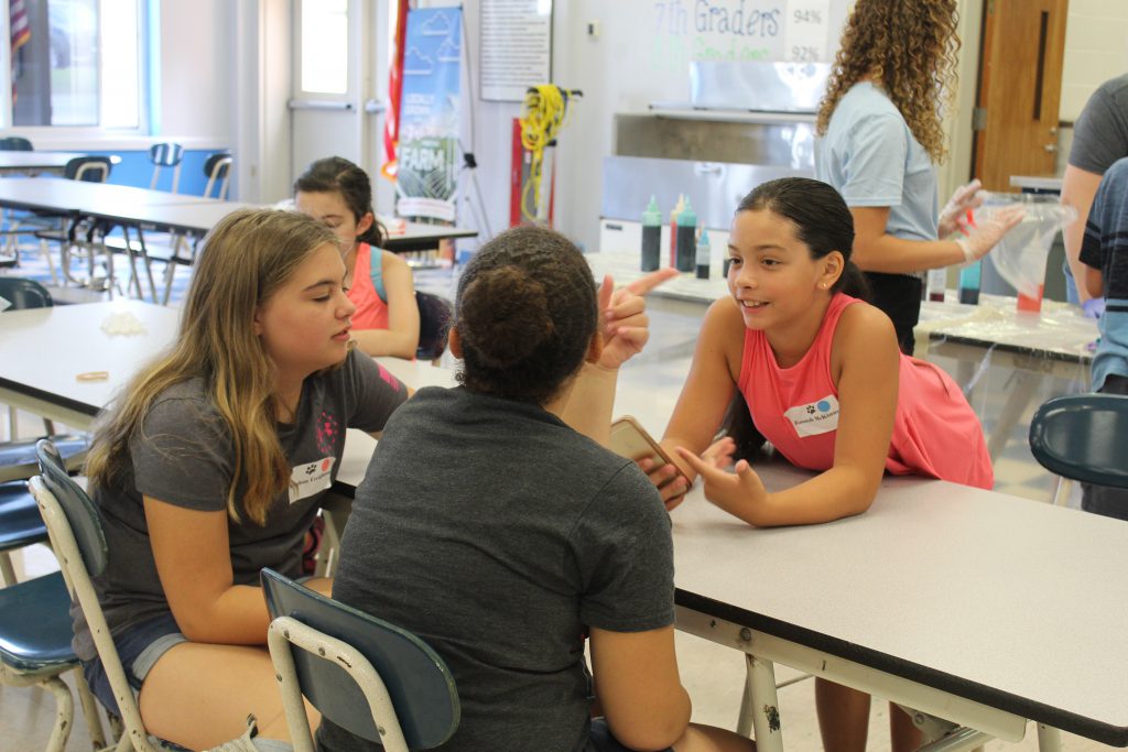 three girls sit at a cafeteria table talking. One is leaning across the table smiling.