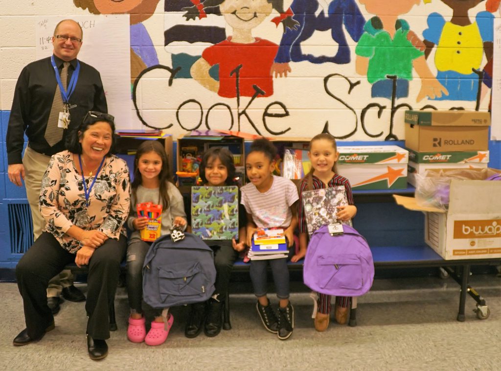 Two adults on the left, one  man standing and a woman sitting, along with four elementary students sitting holding school supplies