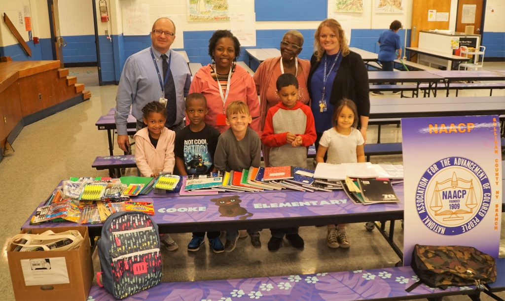 Four adults stand behind five elementary students. In front of them is a table with many school supplies