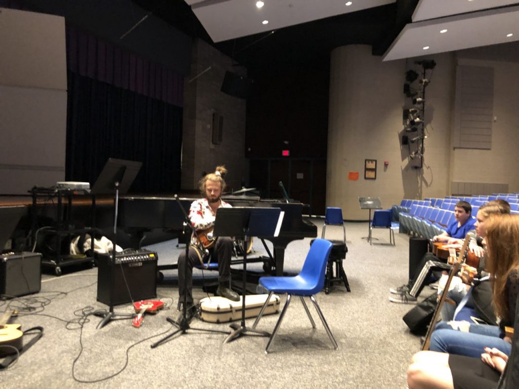 A man sits in front of several students while he plays mandolin. Music stands are all around