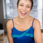 A woman with short dark hair, wearing a dark blue tank with light blue decoration on it smiles broadly.