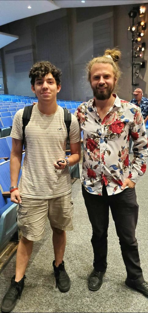 A man in a flowered shirt stands with a high school student in beige clothes wearing a backpack