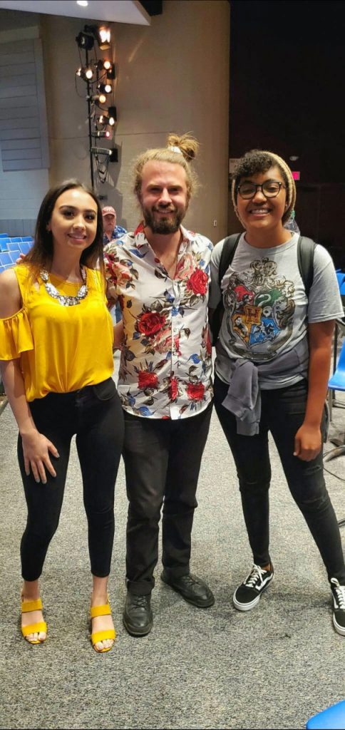 A man with a beard wearing a flowered shirt stands with two high school girls smiling for the camera
