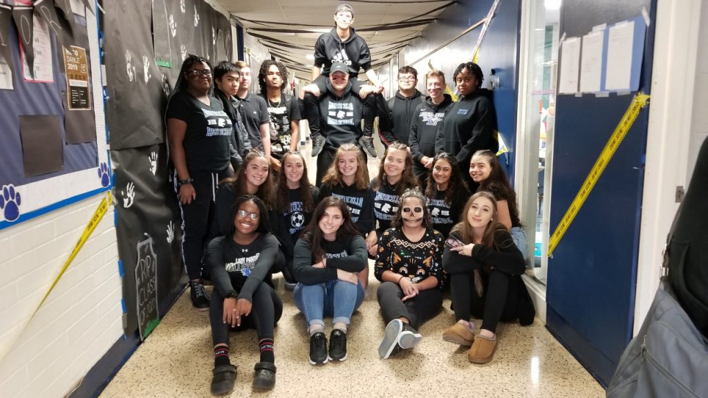 A group of 19 students gather together in a hallway, all wearing Monticello shirts
