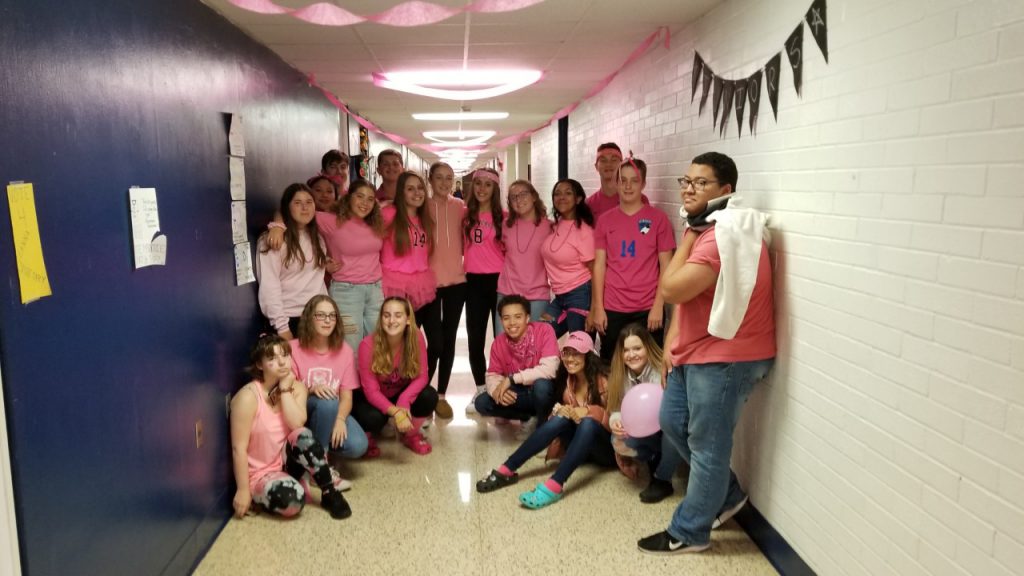 19 high school students stand in a hallway, all wearing pink shirts