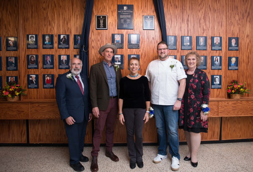 Five adults stand in front of a wall of wood with numerous plaques hanging on it.