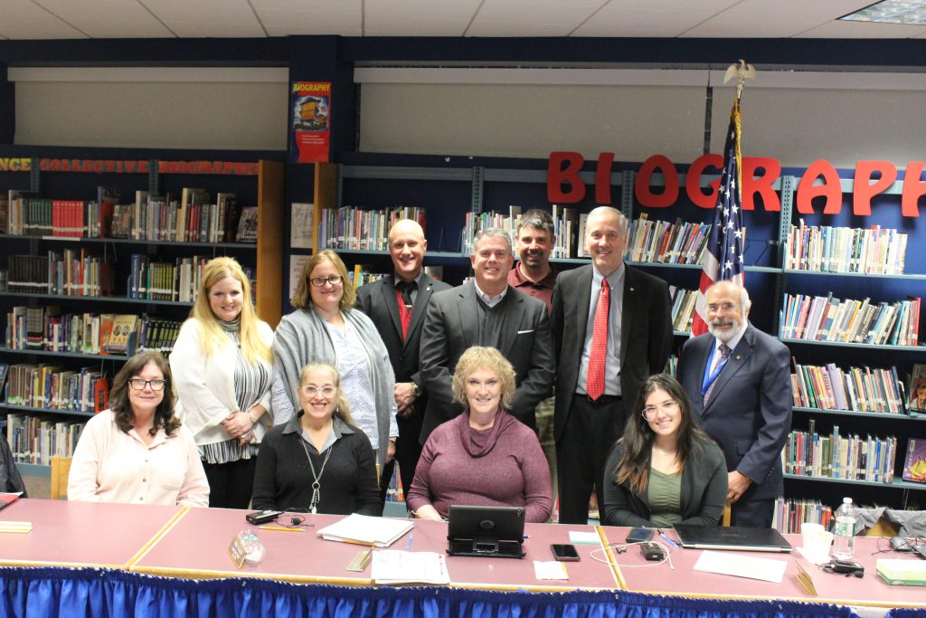 Four women are sitting at a table while two more women and five men stand behind them looking at the camera. Behind them are shelves of books.