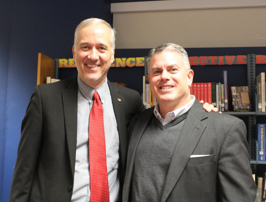 A man with a jacket and red tie stands with his arm around the shoulder of another man. Both are smiling.