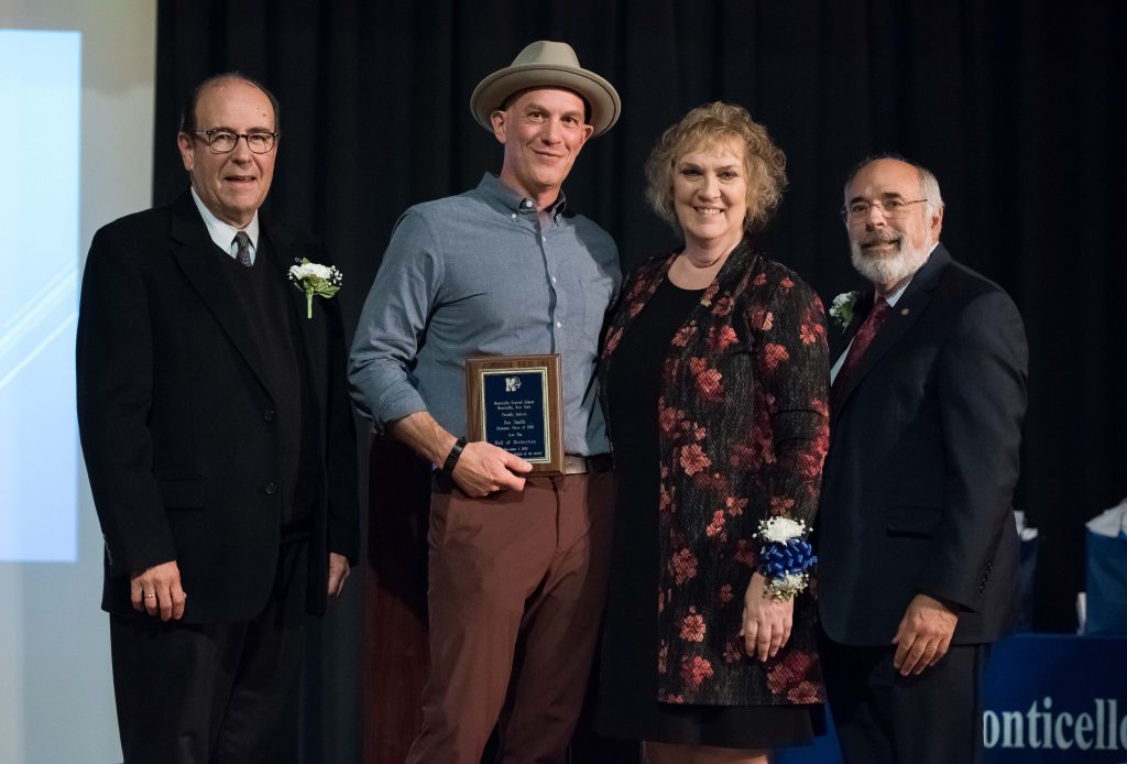 Four adults - three men and one woman - stand together. The man second from left is holding a small plaque he just received. He is wearing a gray hat.