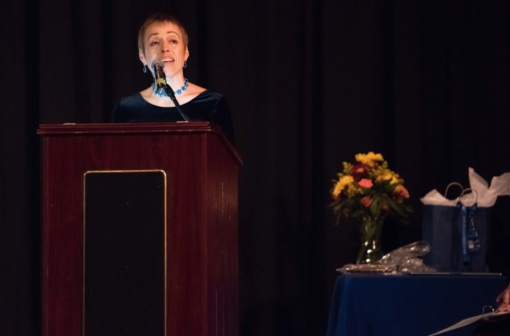 A woman stands at a podium speaking. Next to her is a table with gift bags and a vase filled with autumn flowers.