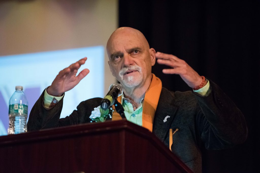 A man with a white goatee wearing an open colored shirt and jacket and orange scarf, talks at a podium. His hands are animated.