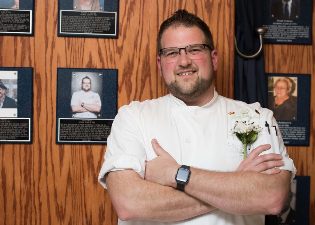 A man in a white chef's jacket stands with his arms crossed in front of his plaque hanging on the wall.