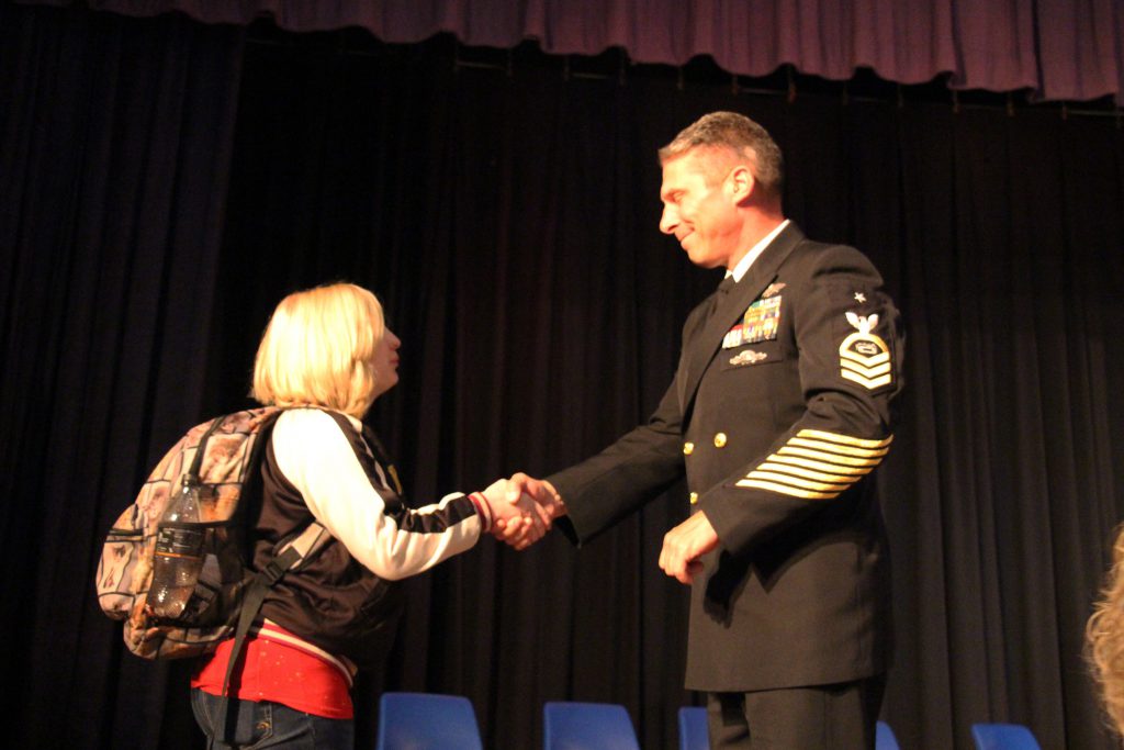 A high school student with blonde hair shakes the hand of a man wearing a uniform.