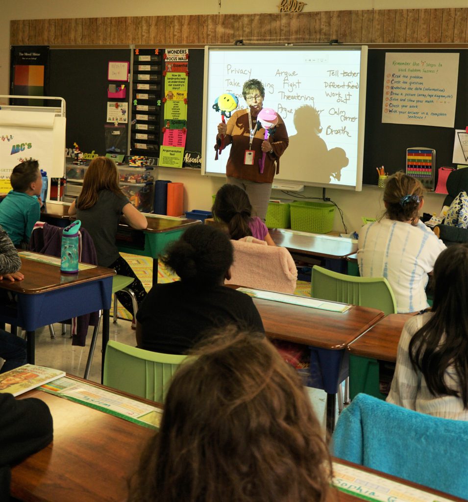 A woman with two puppets is in the front of the class as students sit at their desks, watching and listening to her presentation.