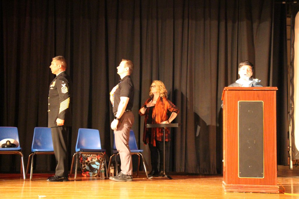 Two men and a woman have their hands over their hears for the pledge of allegiance on a stage