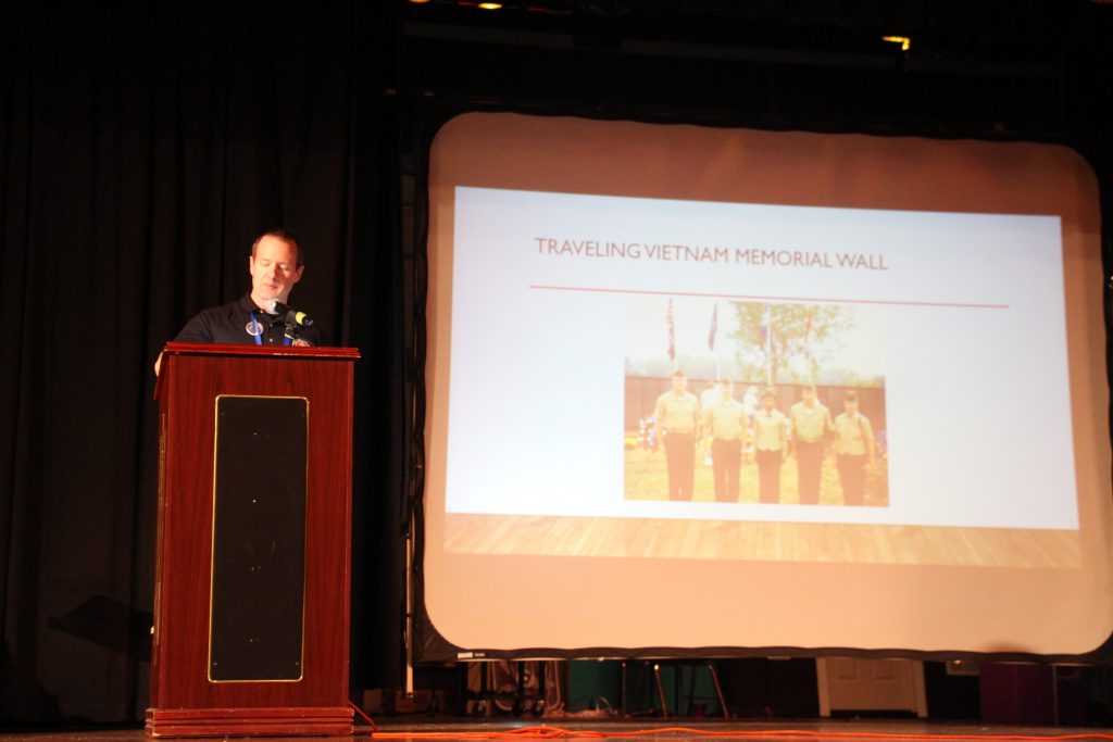 A man at a podium speaks, and there is a picture up on a screen next to him.