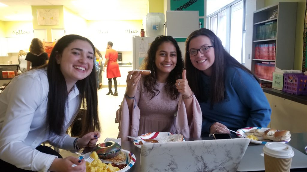 Three high school girls smile while eating breakfast. The one in the middle is giving a thumbs up.