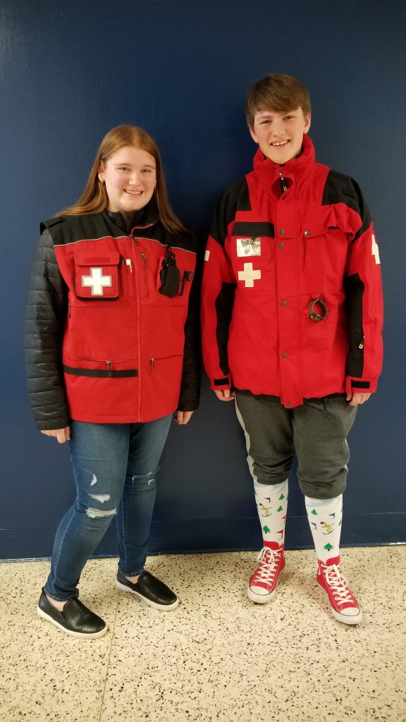 A young woman and young man are dressed in the distinctive red jackets with white crosses on their right side. They are smiling.