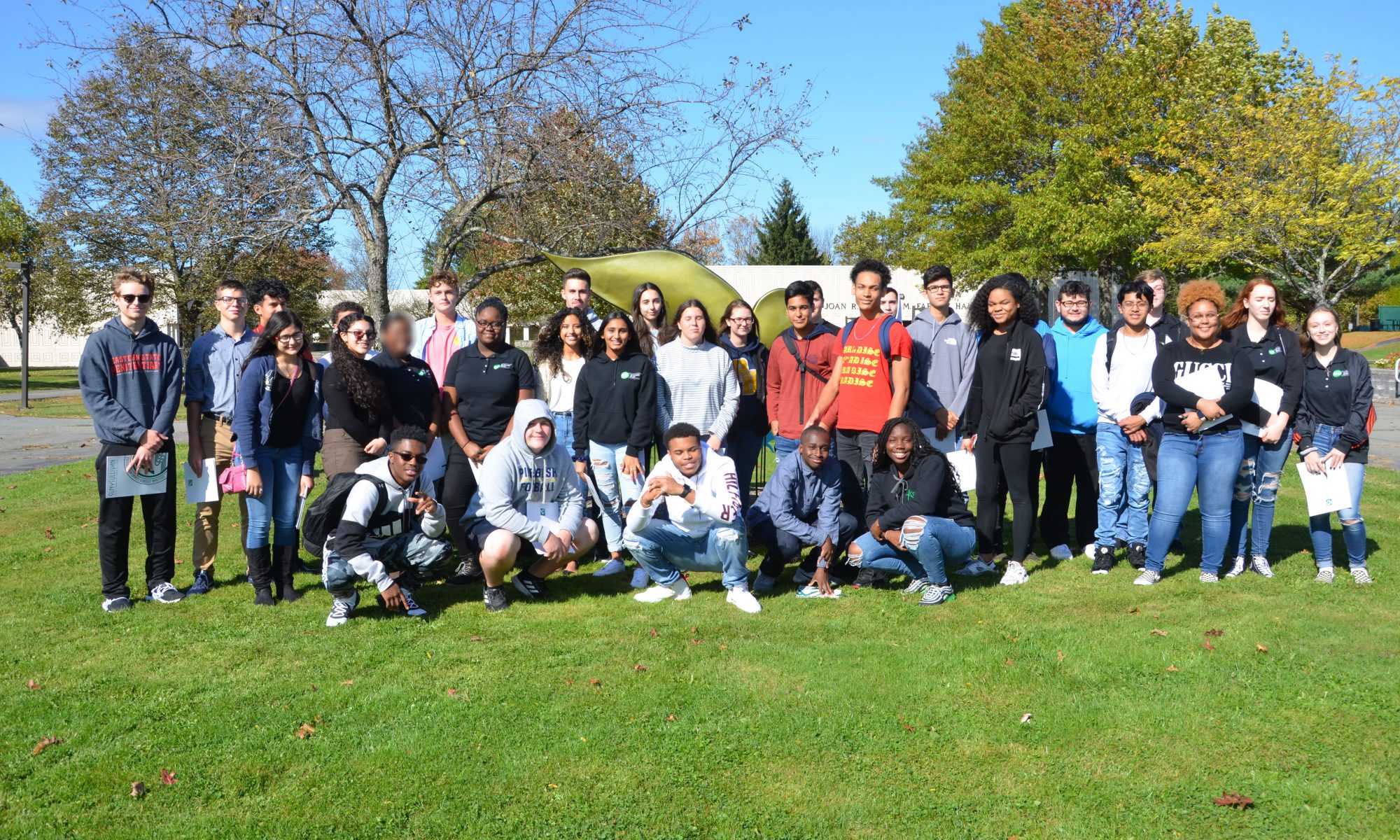 Blue sky, green grass and trees. A large group of students stand and sit for the photo.