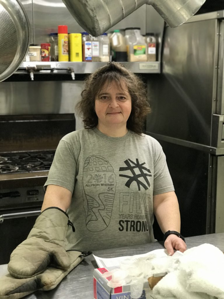 Heidi is in the kitchen wearing an oven mat and smiling