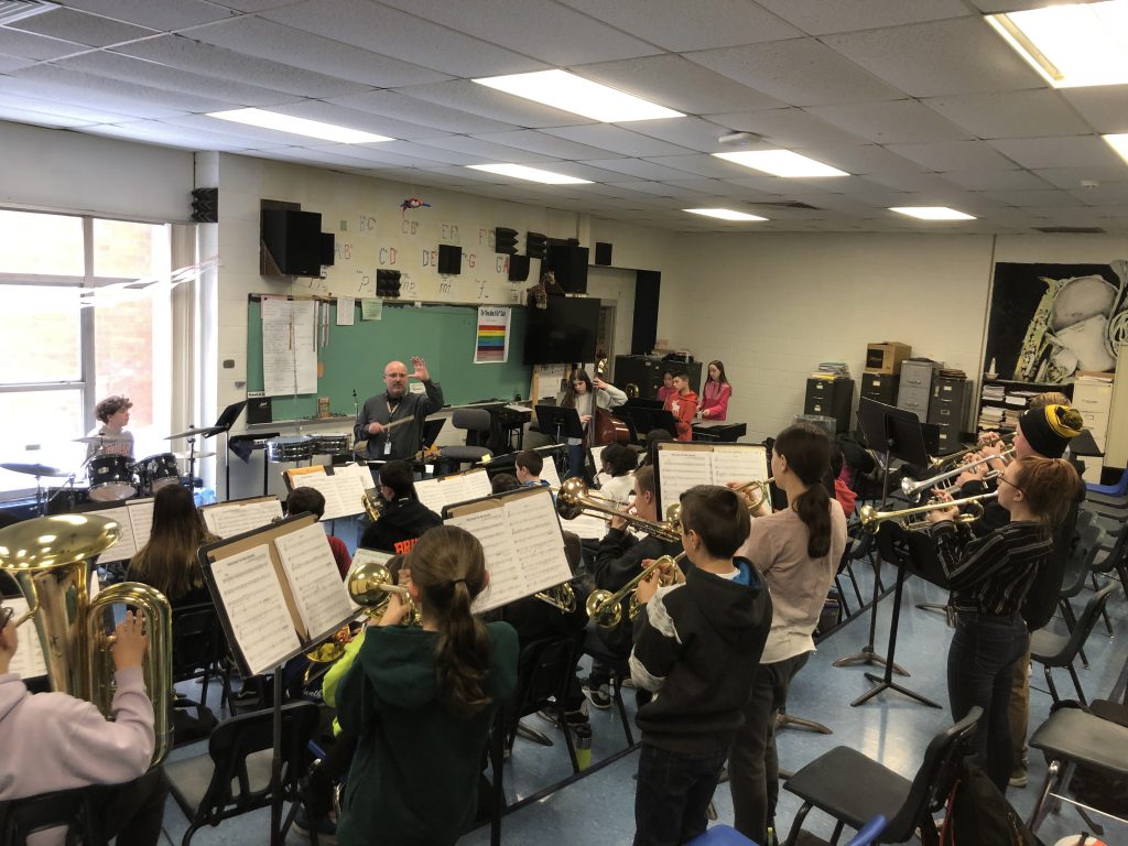 Man stands in front of the room filled with middle school musicians directing them.