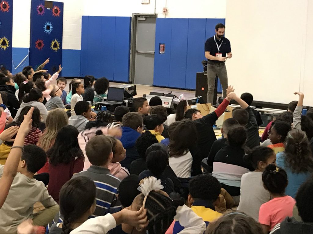 A large group of elementary school students sit facing a screen and a man in a dark shirt and lighter pants.