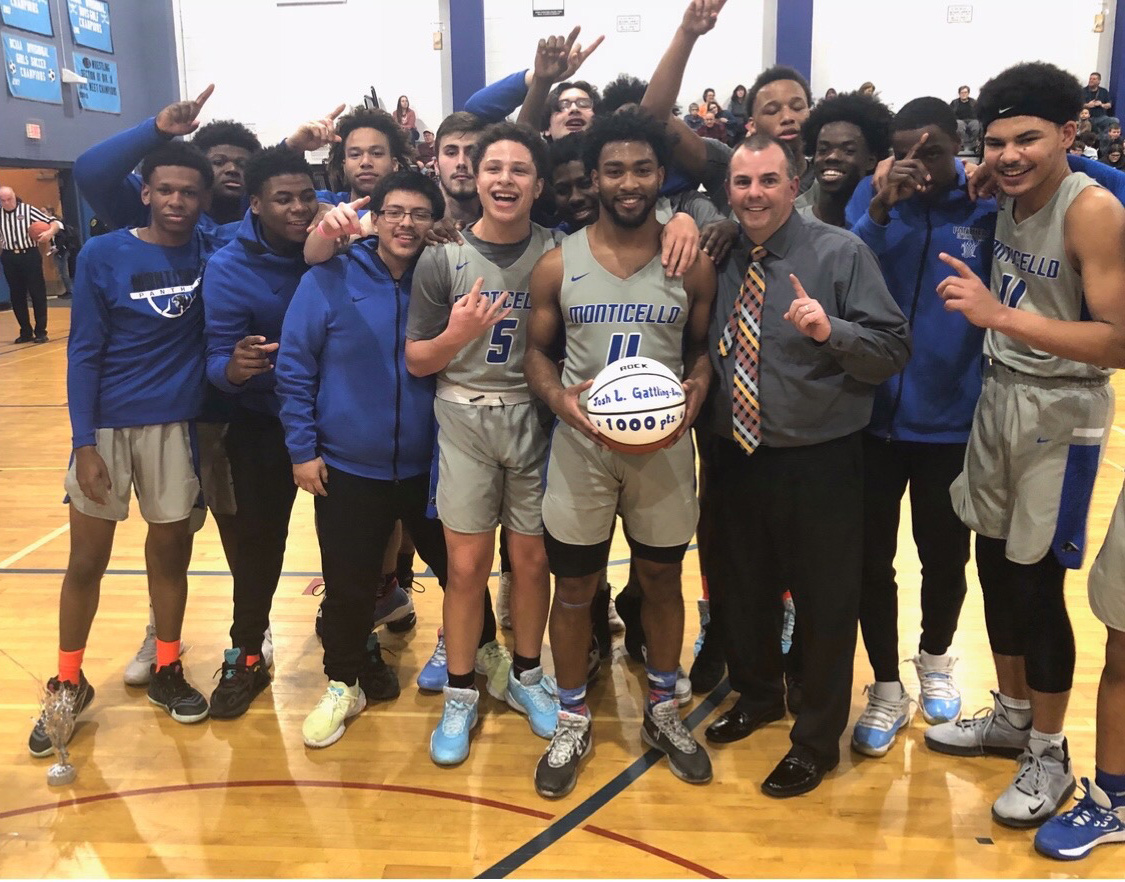 A group of high school basketball players stand around their teammate and coach. The teammate is holding a basketball with his name and 1,000 points written on it.