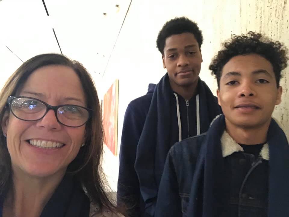 A woman with long brown hair and glasses smiles standing next to two high school students, wearing blue jackets and smiling.