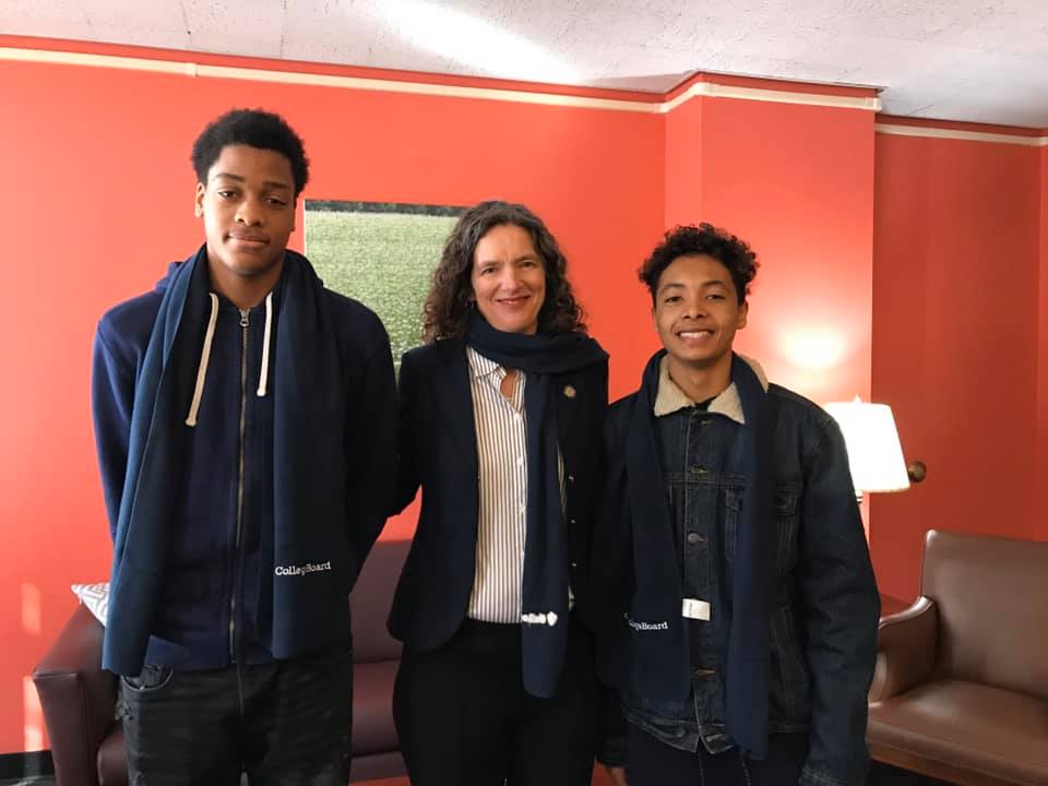 Two high school students stand on either side of a woman in an office. All are smiling.