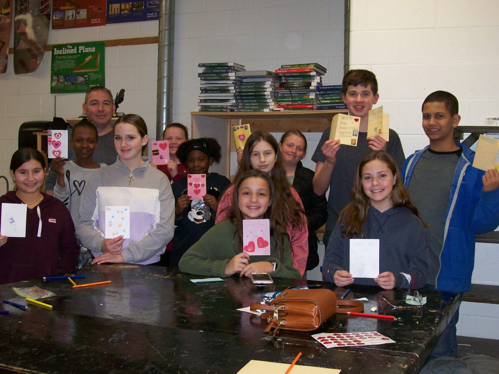 A group of 11 middle school students and one teacher stand holding up the Valentines cards they made