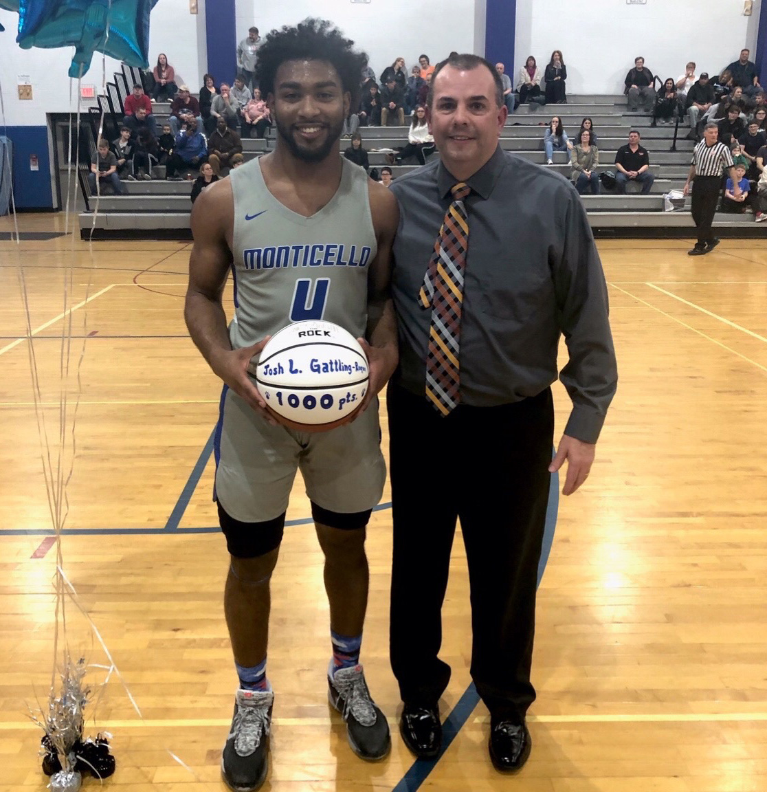 A high school basketball player in a gray and blue uniform holds a basketball painted white with words marking his 1000th point.