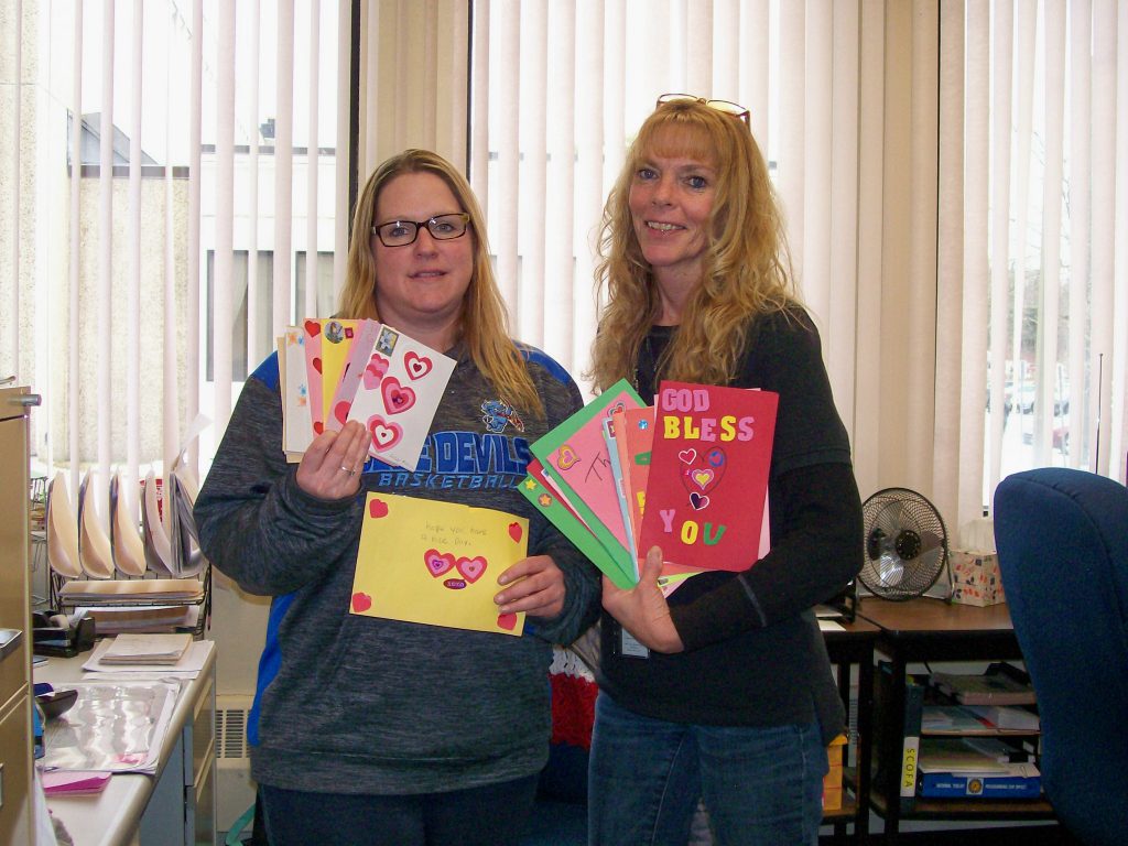 Two women with long blonde hair hold up festiva Valentine's Day cards.