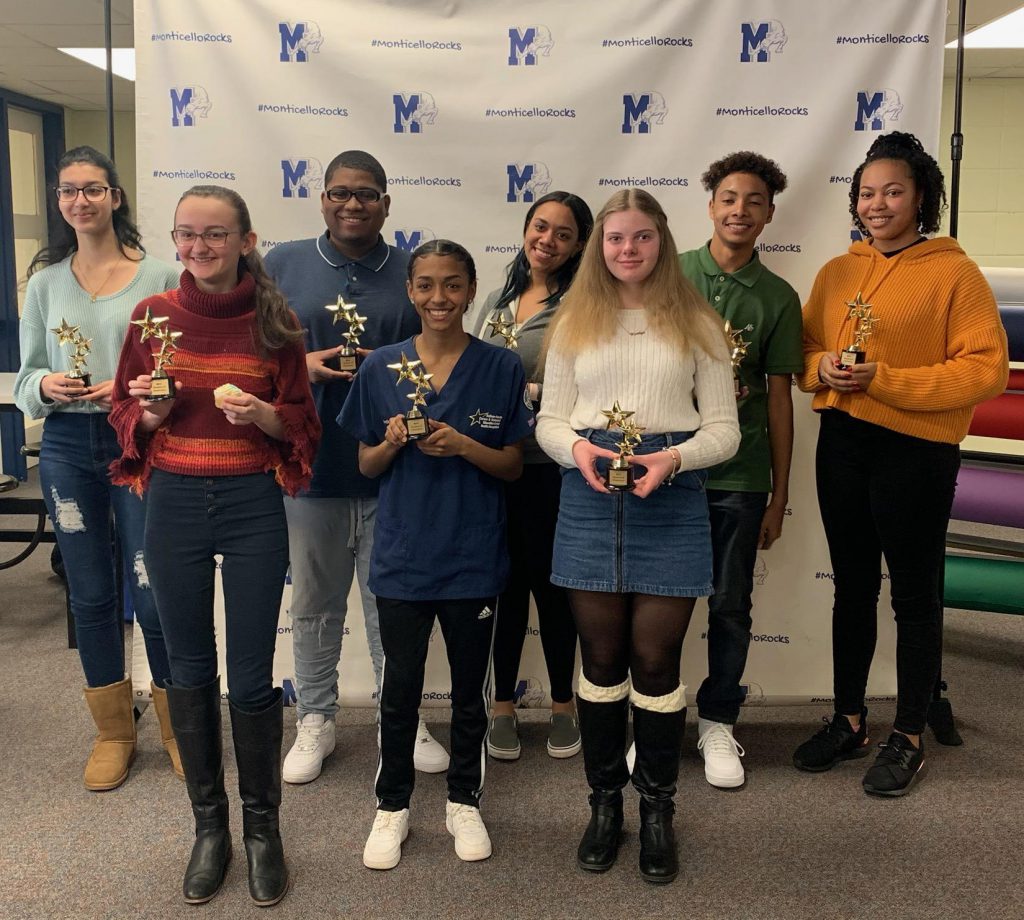 A group of eight high school students holding trophies stand in front of a large white banner with the Monticello logo scattered around it.