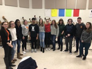 A large group of high school students in a semi-circle smiling. There are instruments behind them.