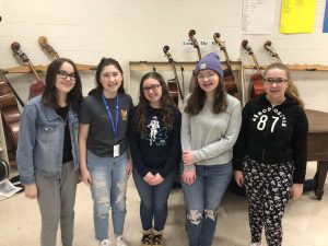 A group of five high school age girls standing in a semi-circle with instruments behind them.