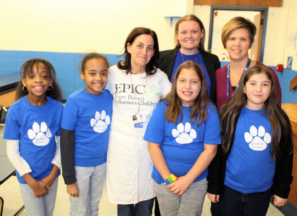 Three adults stand with four older elementary school girls. all of the girls are wearing blue t-shirts with a white panther paw print on them.