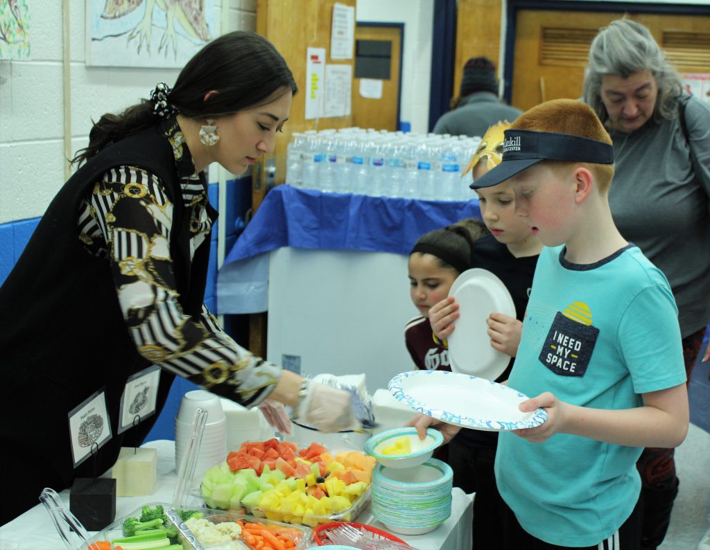 A student wearing a blue shirt and visor holds a plate while a woman wearing a ponytail dishes out fruit to him. The table has lots of fruits and vegetables on it. 