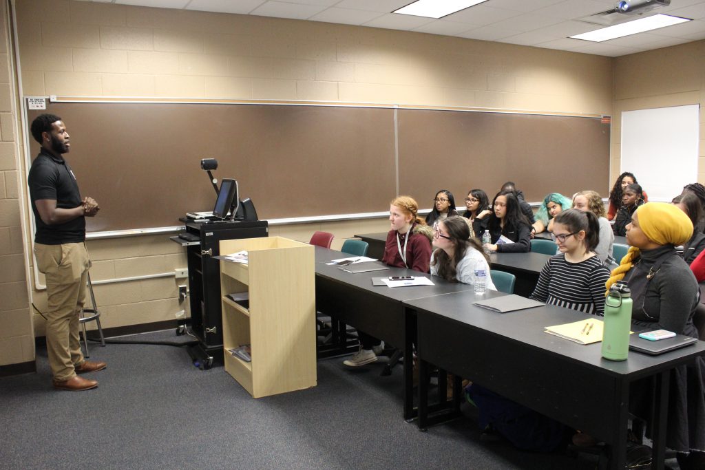 students are seated at desks in a classroom and listening to a teacher 