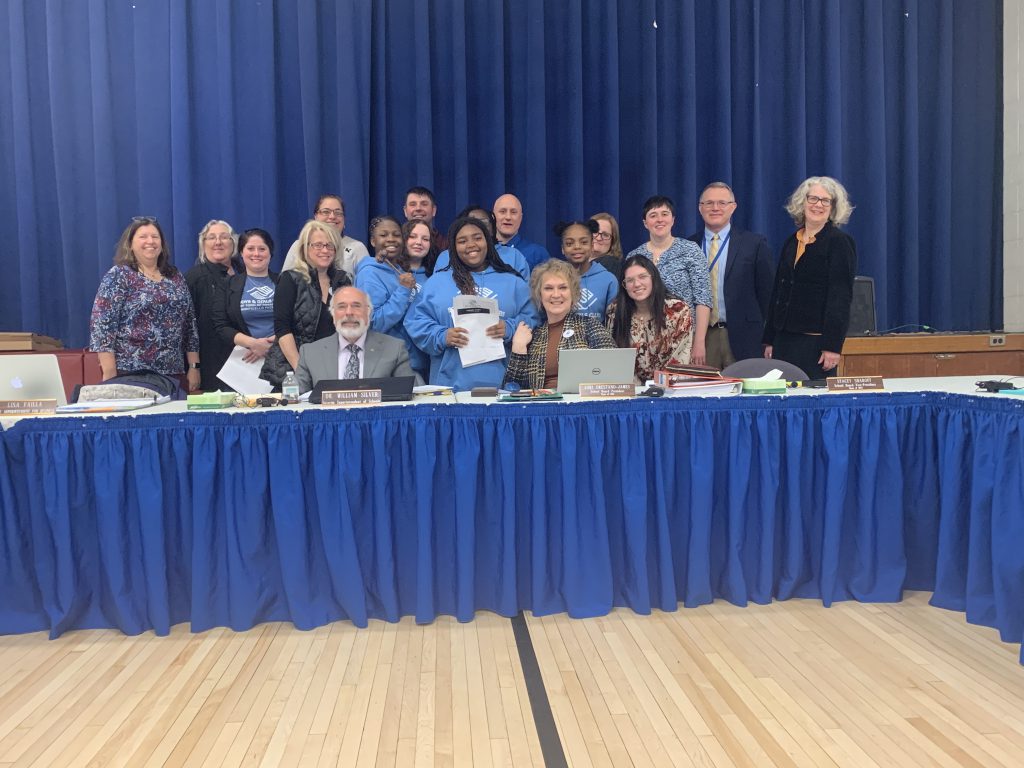 the members of the keystone club are standing behind the Board table with members of the Board of Education. 