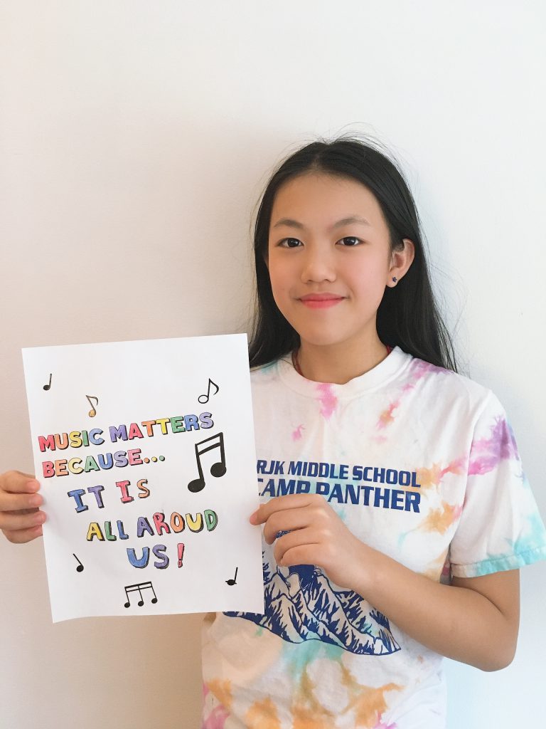student is smiling and holding a piece of paper that says music matters because it is all around us