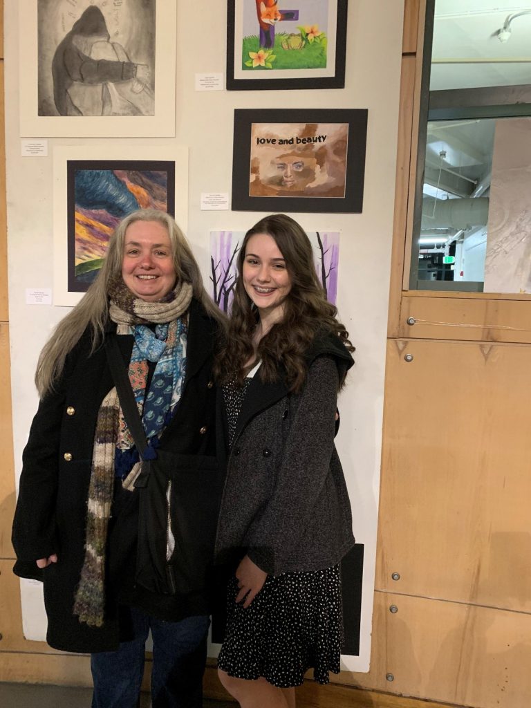 A woman in a dark coat and winter scarf smiles along with a young woman also in a dark coat. They are standing in front of a wall with framed artwork.