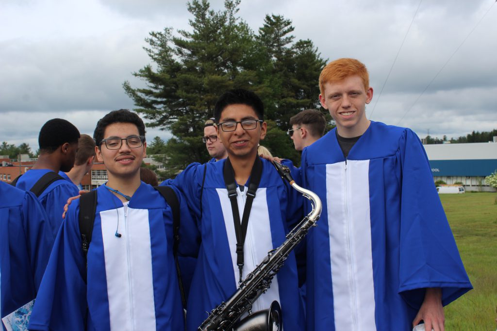 three students in graduation robes are smiling. The student in the center has a saxaphone around his neck