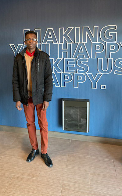 A tall young man wearing a black jacket, yellow shirt and orange pants.